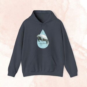 May include: Dark navy blue hoodie with a front pocket and a hood with a drawstring. The front of the hoodie displays a watercolour manatee illustration within a light blue teardrop shape. The text "FLOATY POTATO" is printed below.