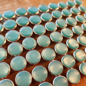 May include: A close-up of a large quantity of teal blue round buttons with silver rims. The buttons are arranged in a grid pattern on a wooden surface.