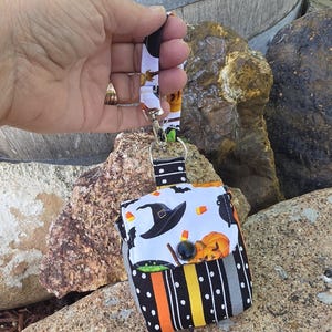 May include: A small fabric pouch featuring a Halloween-themed print with witch hats, pumpkins, and candy corn. The pouch has a flap with a black button closure and a striped pattern of black, orange, yellow, and gray. It includes a fabric wrist strap.