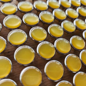 May include: A close-up image of a large collection of yellow glass cabochons, each set in a silver-toned metal setting. The cabochons are arranged in a grid pattern on a wooden surface.