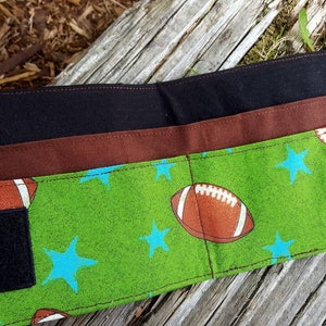 May include: A black and brown fabric wallet with a green and blue fabric flap. The flap has a pattern of stars and footballs. The wallet has two velcro closures.