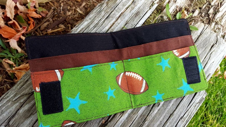 May include: A black and brown fabric wallet with a green and blue fabric flap. The flap has a pattern of stars and footballs. The wallet has two velcro closures.
