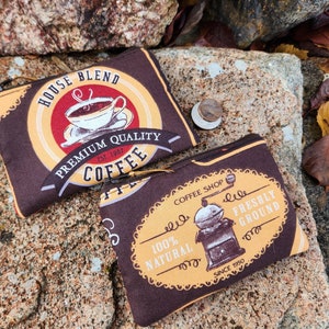 May include: Two brown fabric pouches with zipper closures. The top pouch has a yellow and red design with the text "House Blend, Est. 1997, Premium Quality Coffee". The bottom pouch has a yellow design with the text "Coffee Shop, 100% Natural, Freshly Ground, Since 1950".