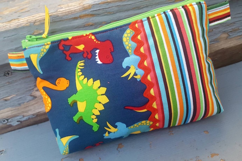 May include: A blue zippered pouch with a colourful striped fabric on one side and a dinosaur print on the other side. The dinosaur print features green, yellow, red, and orange dinosaurs on a dark blue background.
