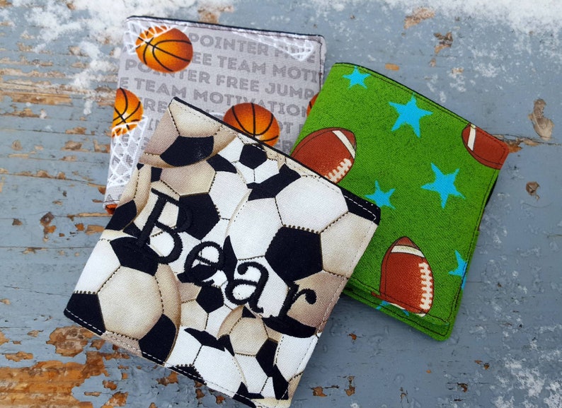 May include: Three fabric wallets with different sports themes. The first wallet has a white background with black and brown soccer balls and the name "Bear" embroidered in black. The second wallet has a white background with a basketball and the words "POINTER FREE TEAM MOTIVATION". The third wallet has a green background with blue stars and brown footballs.