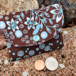 Puede incluir: Una peque&ntilde;a cartera de monedas de tela marr&oacute;n con un patr&oacute;n floral azul y blanco en la solapa delantera y un patr&oacute;n de c&iacute;rculos azul y blanco en la parte posterior. La cartera tiene un cierre de presi&oacute;n y un bot&oacute;n azul claro.