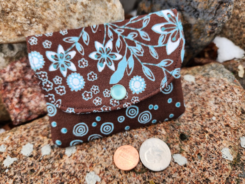 Puede incluir: Una peque&ntilde;a cartera de monedas de tela marr&oacute;n con un patr&oacute;n floral azul y blanco en la solapa delantera y un patr&oacute;n de c&iacute;rculos azul y blanco en la parte posterior. La cartera tiene un cierre de presi&oacute;n y un bot&oacute;n azul claro.