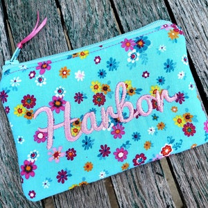 May include: A rectangular zippered pouch in bright turquoise fabric featuring a colorful floral pattern of small red, yellow, and white flowers. The name Harbor is embroidered in pink script across the center, and a pink ribbon pull is attached to the zipper.