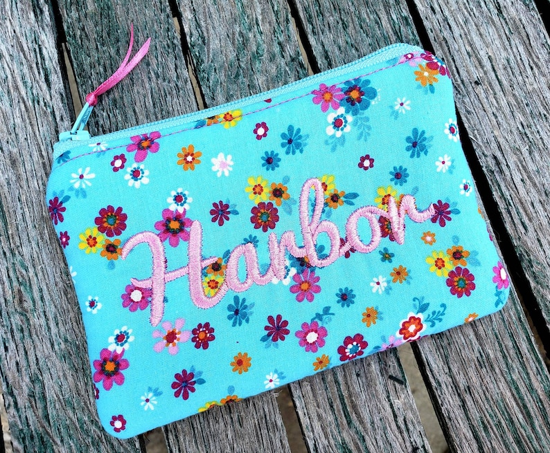 May include: A rectangular zippered pouch in bright turquoise fabric featuring a colorful floral pattern of small red, yellow, and white flowers. The name Harbor is embroidered in pink script across the center, and a pink ribbon pull is attached to the zipper.