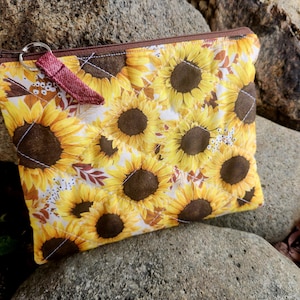 May include: A rectangular, zippered pouch featuring a vibrant sunflower pattern in yellow, brown, and white. The pouch has a brown zipper and a maroon fabric loop attached to a silver key ring. The pouch is resting on a bed of gray rocks.
