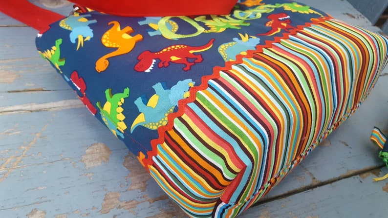 May include: A blue tote bag with a dinosaur print and a colourful striped bottom. The bag has a red scalloped trim.
