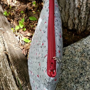 K&ouml;nnte beinhalten: Eine graue Rei&szlig;verschlusstasche mit Blumendruck und einem roten Rei&szlig;verschluss. Die Tasche liegt auf einer Holzoberfl&auml;che.