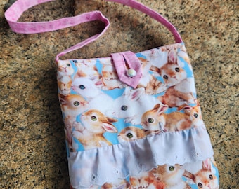 May include: A small blue and white fabric bag with a pink strap. The bag has a white ruffle trim and is covered in a pattern of bunnies.