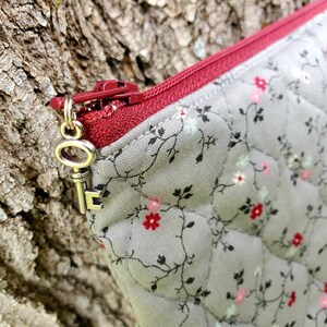 K&ouml;nnte beinhalten: Eine graue Rei&szlig;verschlusstasche mit einem roten Rei&szlig;verschluss und einem silbernen Schl&uuml;sselanh&auml;nger. Die Tasche hat ein Blumenmuster mit kleinen rosa, wei&szlig;en und blauen Bl&uuml;ten.