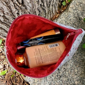 K&ouml;nnte beinhalten: Eine graue und wei&szlig;e, florale Rei&szlig;verschlusstasche mit rotem Futter. Die Tasche ist offen und enth&auml;lt eine Tube Handcreme, eine Tube Mascara und eine kleine Flasche Lotion.