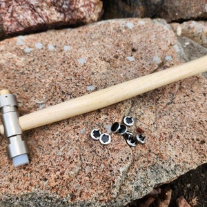 May include: A wooden-handled hammer with a metal head and a rubber base. The hammer is lying on a rock surface with a pile of small, round, silver-colored metal pieces with black centers.