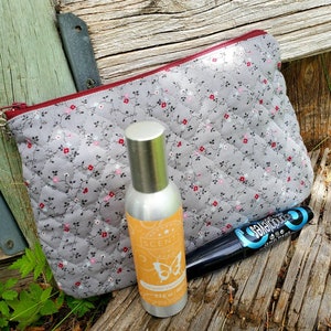 K&ouml;nnte beinhalten: Eine graue, gesteppte Kosmetiktasche mit Rei&szlig;verschluss und Blumenmuster, eine silberne Spr&uuml;hflasche mit gelbem Etikett, auf dem "SCENTSY Caramel Vanilla 2.4 fl oz / 71 ml" steht, und eine schwarze Mascara mit blauer Aufschrift "BELLISSIMA" auf einer Holzoberfl&auml;che.