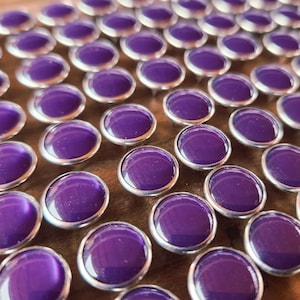 May include: Close-up of numerous purple cabochons with silver-colored metal settings. The oval-shaped stones have a glossy finish and are arranged closely together. The cabochons are likely for jewelry making or craft projects.