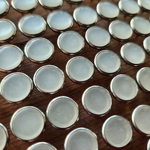 May include: A close-up of a large collection of white, round, decorative push pins with silver metal rims. The push pins are arranged in rows on a wooden surface.