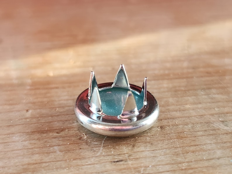 Puede incluir: Un bot&oacute;n de metal plateado con cinco puntas afiladas, dise&ntilde;ado para sujetar tela. El bot&oacute;n tiene un centro azul verdoso.