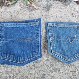 Pu&ograve; includere: Due tasche di jeans in denim blu. Una tasca ha una cucitura orizzontale al centro. L'altra tasca ha un motivo di cucitura decorativo color oro. Entrambe le tasche sono su una superficie di pietra grigia.