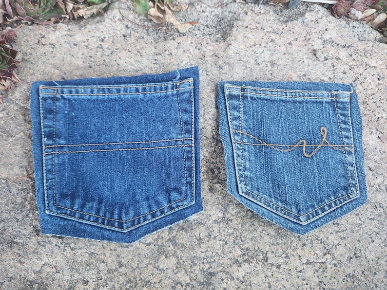 Pu&ograve; includere: Due tasche di jeans in denim blu. Una tasca ha una cucitura orizzontale al centro. L'altra tasca ha un motivo di cucitura decorativo color oro. Entrambe le tasche sono su una superficie di pietra grigia.