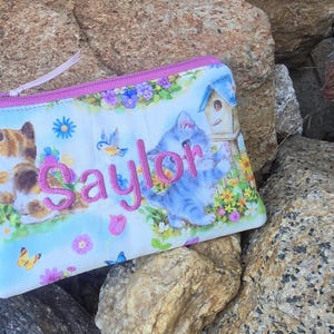 May include: A rectangular zippered pouch featuring a light blue background with a colorful print of kittens, flowers, butterflies, and a birdhouse. The name Saylor is embroidered in pink thread across the center of the pouch.
