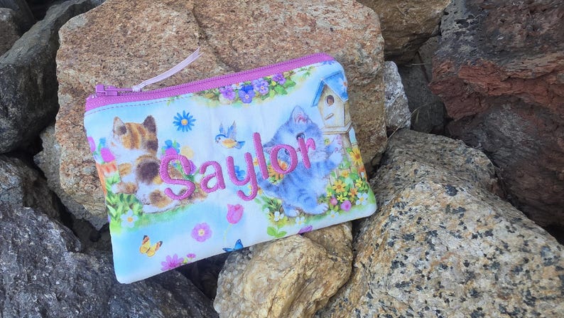 May include: A rectangular zippered pouch featuring a light blue background with a colorful print of kittens, flowers, butterflies, and a birdhouse. The name Saylor is embroidered in pink thread across the center of the pouch.