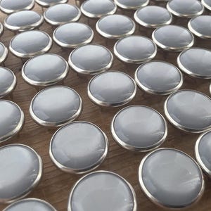 May include: A large collection of circular upholstery tacks arranged in rows. Each tack features a smooth, light gray, domed top surface encased in a polished silver-toned metal rim, creating a uniform and repetitive pattern across the display.
