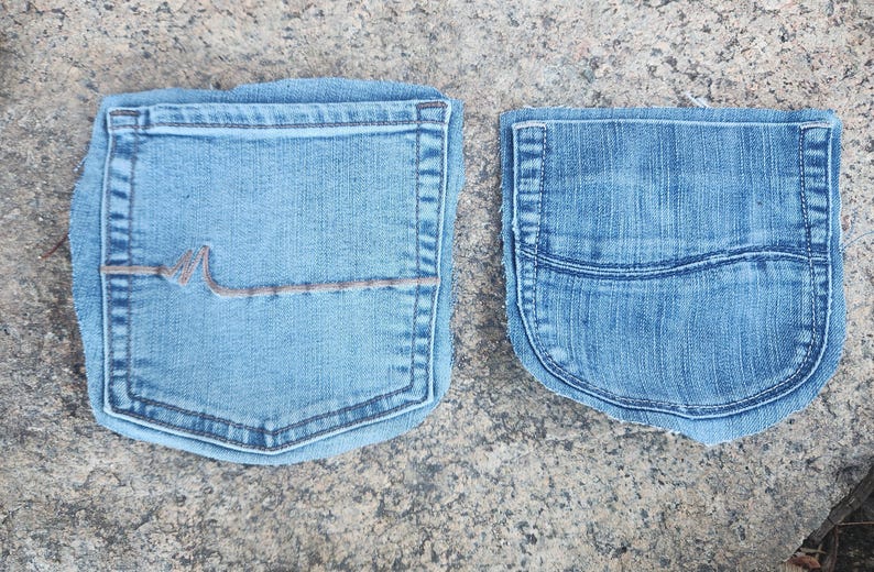 Pu&ograve; includere: Due tasche di jeans in denim blu chiaro. Una tasca ha un design decorativo con cuciture marroni. L'altra tasca ha una forma arrotondata con una cucitura curva. Entrambe le tasche sono su una superficie grigia strutturata.