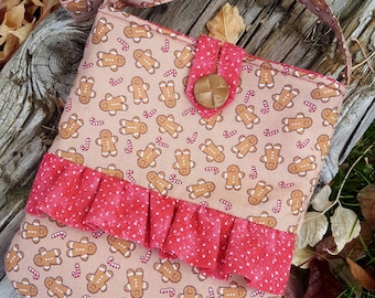 May include: A beige crossbody bag with a gingerbread man and candy cane pattern. The bag features a red ruffled accent and a wooden button closure. The strap and flap match the gingerbread pattern.