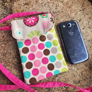 May include: A pink and white polka dot phone case with a flap closure and a pink strap. The case is made of fabric and has a brown corduroy texture. The phone case is lying on a brown and gray textured surface. The text "GALAXY S III" is visible on the back of a black phone.