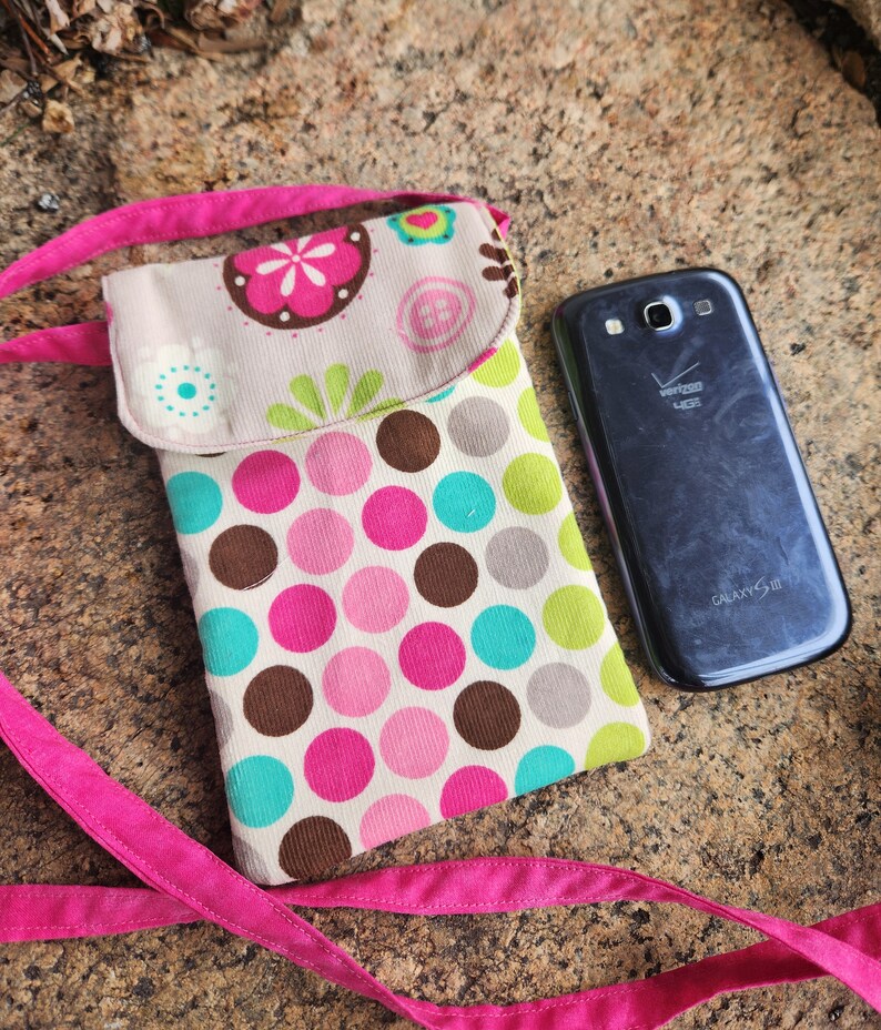 May include: A pink and white polka dot phone case with a flap closure and a pink strap. The case is made of fabric and has a brown corduroy texture. The phone case is lying on a brown and gray textured surface. The text "GALAXY S III" is visible on the back of a black phone.