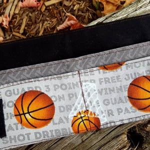 May include: A black and gray fabric wallet with a basketball theme. The wallet has a zipper closure and features a variety of basketball-related words and images, including "Free Jump", "3 Pointers", and "Team Motivated".