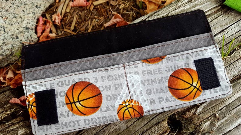 May include: A black and gray fabric wallet with a basketball theme. The wallet has a zipper closure and features a variety of basketball-related words and images, including "Free Jump", "3 Pointers", and "Team Motivated".