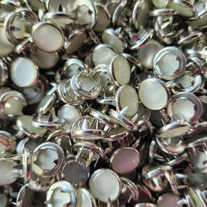 May include: A close-up of a pile of silver-toned metal buttons with white, pearlescent centers. The buttons have a decorative, pronged design on the back.