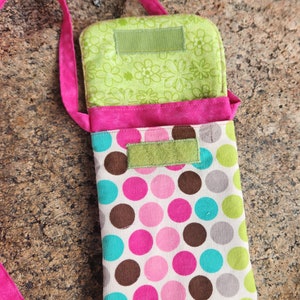 May include: A pink and green polka dot fabric phone pouch with a pink strap. The pouch has two green fabric flaps with velcro closures.
