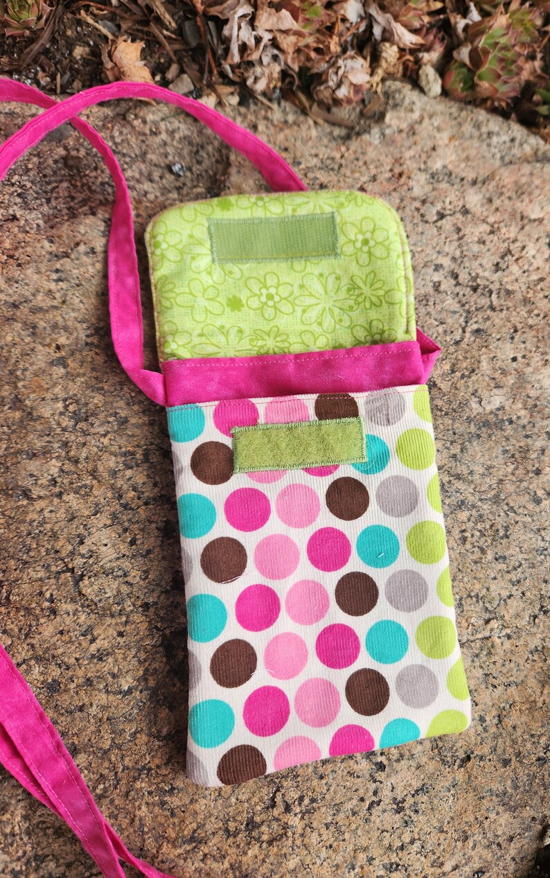 May include: A pink and green polka dot fabric phone pouch with a pink strap. The pouch has two green fabric flaps with velcro closures.