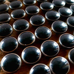 May include: A close-up of a collection of black round buttons with a silver metal rim. The buttons are arranged in rows on a wooden surface.