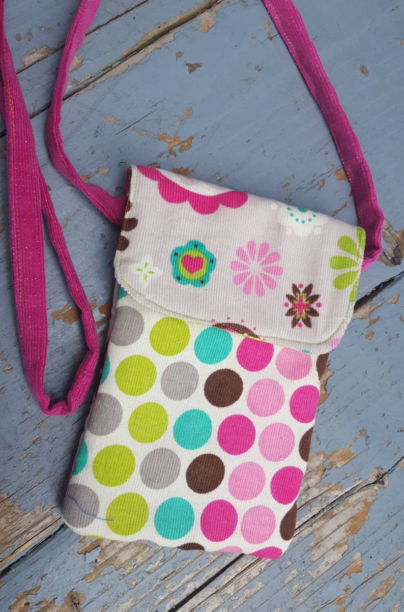 May include: A small pink and white fabric phone case with a polka dot pattern and floral design. The case has a pink strap.