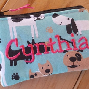 May include: A rectangular fabric zipper pouch featuring a light blue background with a repeating pattern of cartoon dogs and paw prints. The name Cynthia is embroidered in bright pink thread across the center. A black zipper with a pink pull tab.