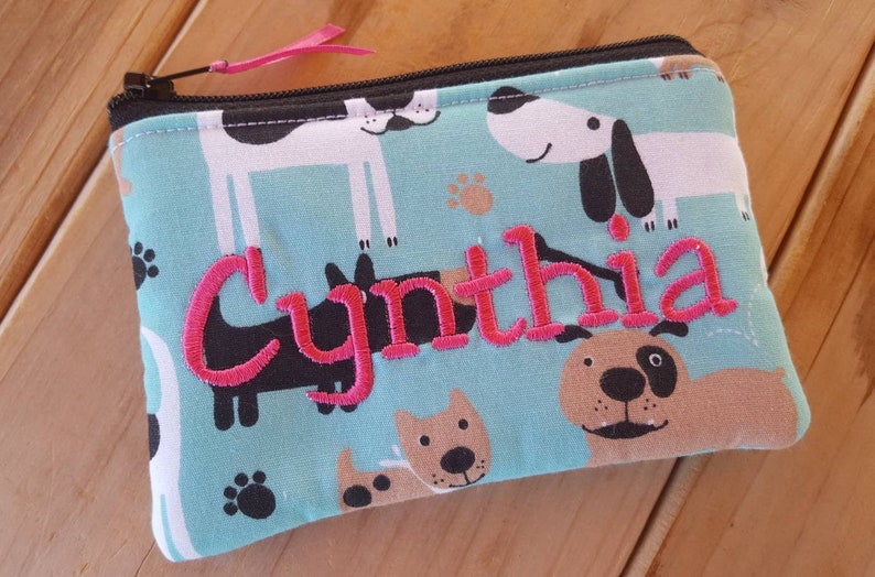 May include: A rectangular fabric zipper pouch featuring a light blue background with a repeating pattern of cartoon dogs and paw prints. The name Cynthia is embroidered in bright pink thread across the center. A black zipper with a pink pull tab.