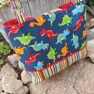 May include: A blue tote bag with a colourful dinosaur print. The bag has a red lining and a striped trim. The bag has two straps.