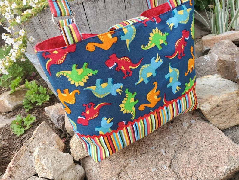 May include: A blue tote bag with a colourful dinosaur print. The bag has a red lining and a striped trim. The bag has two straps.