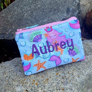 May include: A light blue zippered pouch featuring a repeating pattern of pink mermaids, orange starfish, and purple seashells with bubbles. The name Aubrey is embroidered in dark purple thread across the center of the pouch with a pink zipper.