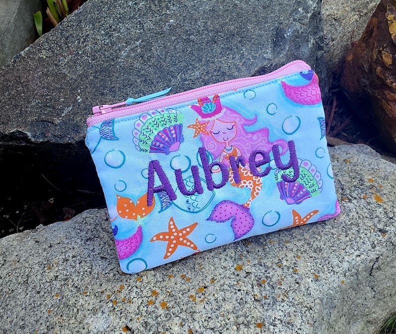 May include: A light blue zippered pouch featuring a repeating pattern of pink mermaids, orange starfish, and purple seashells with bubbles. The name Aubrey is embroidered in dark purple thread across the center of the pouch with a pink zipper.