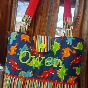 May include: A blue and green tote bag with a dinosaur print and the name "Owen" embroidered in green. The bag has red straps and a striped pocket.