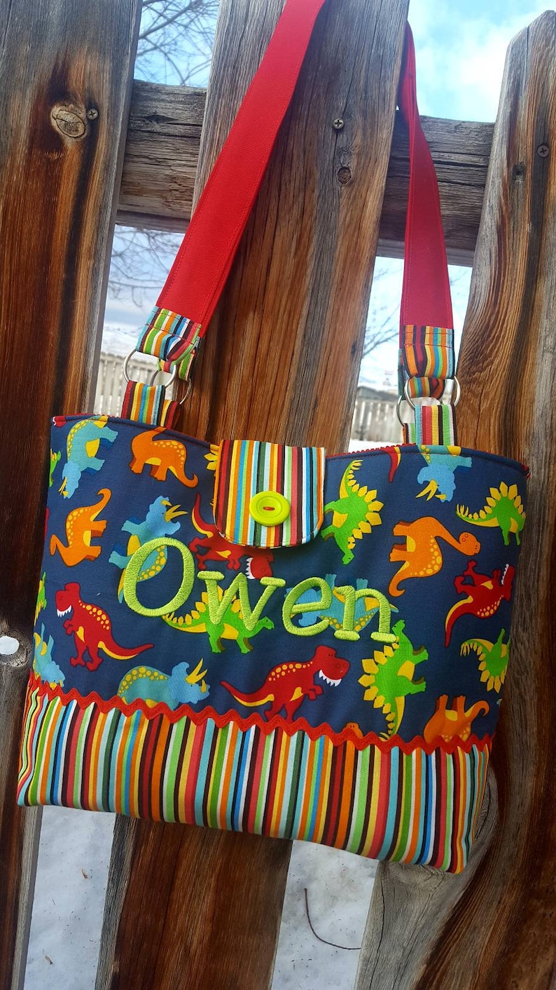 May include: A blue and green tote bag with a dinosaur print and the name "Owen" embroidered in green. The bag has red straps and a striped pocket.