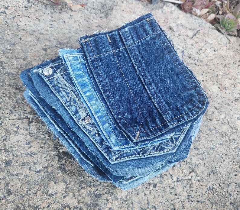 Pu&ograve; includere: Una pila di tasche in denim in varie tonalit&agrave; di blu. Alcune tasche hanno cuciture decorative e dettagli di strass. Le tasche sono disposte su una superficie grigia strutturata.