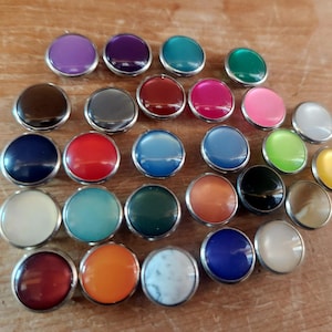 May include: A collection of colorful, round buttons with silver rims. The buttons are in various shades, including purple, teal, green, pink, and blue. They are arranged on a light brown wooden surface.